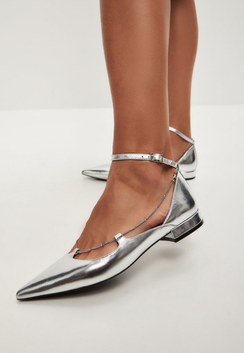 Metallic silberne flache Schuhe mit spitzem Toe, Knöchelriemen und Kettendetails. Glatte Textur mit niedrigem Absatz und klarem Design.