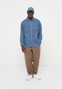 Denim shirt in light blue with two chest pockets and logo patch, paired with brown pants featuring a spotted pattern and white sneakers.