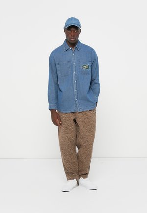 Lichtblauw spijkerblouse met twee borstzakken en logo-opdruk, gecombineerd met bruine broek met een gespikkeld patroon en witte sneakers.