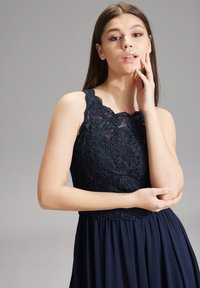 Marineblaues Kleid mit Spitzenoberteil und floralem Stickmuster, ärmelloses Design und plissierter Rock, der eine glatte Stofftextur zeigt.