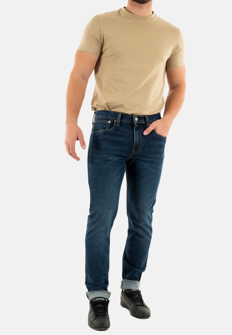 T-shirt beige attillato e jeans in denim blu scuro, con risvolti, abbinati a sneakers nere. Tessuto liscio, stile casual, senza motivi visibili.