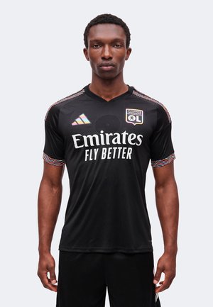 Man standing facing forward wearing black Olympique Lyonnais soccer jersey with "Emirates Fly Better" sponsor and colorful trim on sleeves.