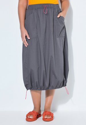 Donna con gonna grigia al ginocchio con tasche e orlo elastico, maglia arancione e sandali rossi intrecciati, in piedi su sfondo bianco.