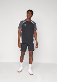 Zwarte sport t-shirt met grijze accenten, korte mouwen en logo. Gecombineerd met bijpassende shorts. Model draagt witte sneakers en sokken.
