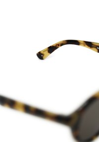 Silmukat aurinkolasit, joissa on pyöristetty kehys, tummat linssit ja teksturoituneet templeet. Lasit esittävät ruskean ja kermanvärin yhdistelmää.