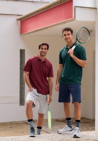 Dva muži v polo tričkách a pruhovaných ponožkách stojí se tenisovými raketami před budovou s červeným přístřeškem.
