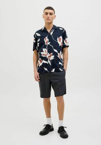 Camisa de manga corta en azul marino con patrones florales en blanco y naranja, combinada con pantalones cortos oscuros. El modelo lleva zapatos negros y calcetines blancos.