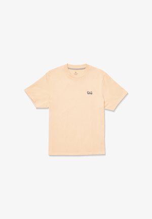 Camiseta de algodón color durazno con mangas cortas, cuello redondo y un pequeño parche de logo negro en el lado izquierdo del pecho. Textura suave y corte relajado.