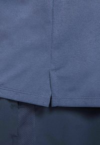 Blaues Sportoberteil aus strukturiertem, atmungsaktivem Stoff. Verfügt über seitliche Schlitze und abgesteppten Saum für eine lockere Passform, kombiniert mit dunkelblauer Hose.