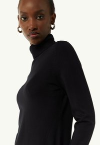 Pull en col roulé noir en tissu tricoté lisse, présentant une coupe ajustée et des détails côtelés au niveau du col et des poignets.