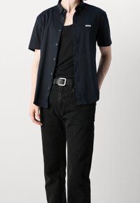 Marineblauw shirt met korte mouwen en kraag, knoopsluiting aan de voorkant en wit logo, gecombineerd met zwarte jeans en een zwarte riem met een zilveren gesp.