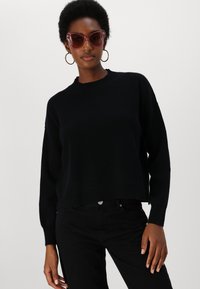 Svart strikket genser med rund hals og senkede skuldre, kombinert med svarte bukser. Tilbehøret inkluderer rosa oversized solbriller og hoops øreringer.