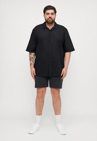 Svarte kortermede skjorter med knapper laget av lett stoff, kombinert med mørk denimshorts. Modellen har på seg hvite joggesko og ankelsokker.