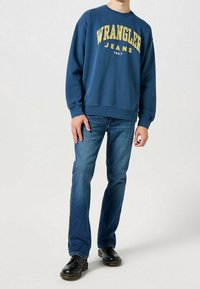 Sweat-shirt bleu avec texte jaune "Wrangler Jeans", associé à un jean bleu foncé et des chaussures noires. La tenue présente une coupe décontractée.