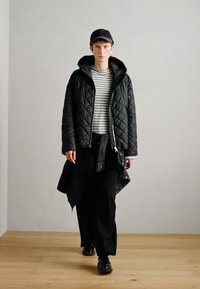 Izšūta melna jaka, zem tās melna svītraina krekls, melnas platās bikses un melnas kurpes. Modelis valkā melnu cepuri, demonstrējot ikdienišķu izskatu.