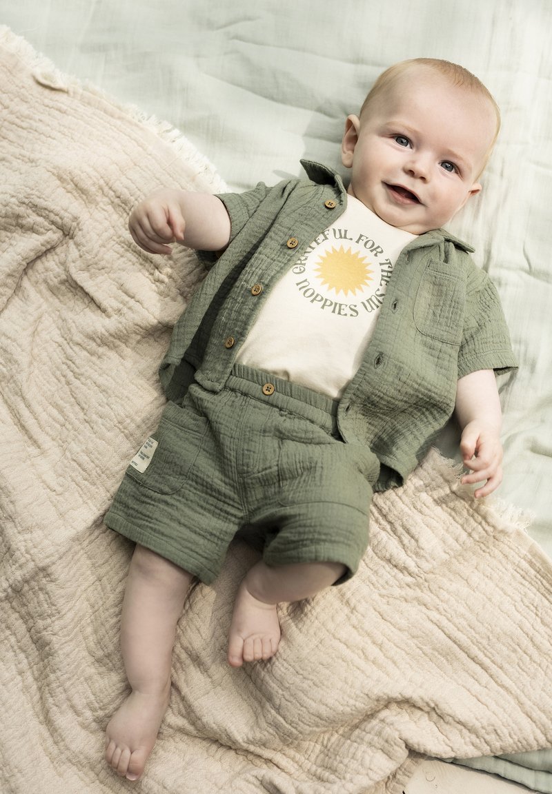 Groene katoenen set van een T-shirt met korte mouwen en korte broek, met een crèmekleurig T-shirt met zonafbeelding. Zachte, gestructureerde stof. Baby ligt op een neutrale deken.