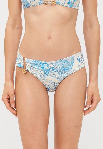 Donna che indossa un bikini bianco e blu con motivi a tema marino e charm dorati circolari sul fianco sinistro.