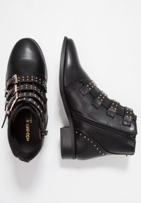 Bottes de cheville noires en simili cuir lisse, présentant plusieurs sangles avec des clous dorés, un talon incurvé et une fermeture éclair sur le côté pour un enfilage facile.