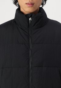 Chaqueta negra acolchada con cuello alto, cierre frontal con cremallera y tejido texturizado. Presenta un diseño acolchado y no tiene logotipos o patrones visibles.