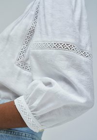 Blusa blanca de tejido texturizado, con intrincados bordes de encaje en los puños y el cuello, complementada con mangas abullonadas.