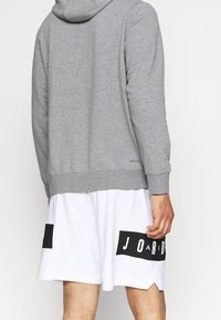 Hombre vistiendo una sudadera con capucha gris de manga larga y pantalones cortos de baloncesto blancos con paneles del logo negro de Jordan en la parte trasera.