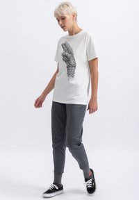 Witte crewneck t-shirt met een gedetailleerde graphic van een hand, gecombineerd met grijze taps toelopende broek en zwarte sneakers met witte veters.