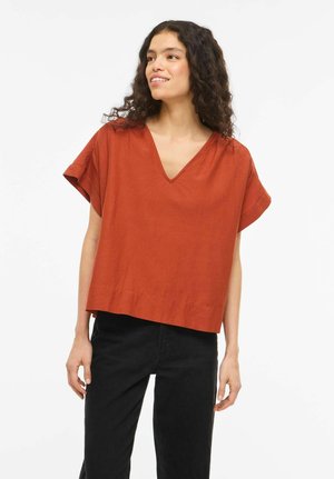 Kvinde med krøllet hår iført en løs rust-orange T-shirt med V-hals og korte ærmer samt sorte bukser, der kigger let til siden og smiler.