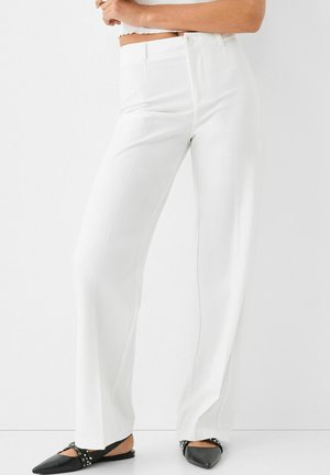 Kobieta ubrana w białe, szerokie spodnie z wysokim stanem oraz czarne, szpiczaste baleriny z srebrnymi ćwiekami, stojąca na tle jednolitej białej ściany.