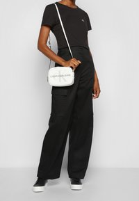 Osoba v černém tričku a širokých černých kalhotách, držící bílou crossbody tašku Calvin Klein Jeans, stojí na jednobarevném pozadí.