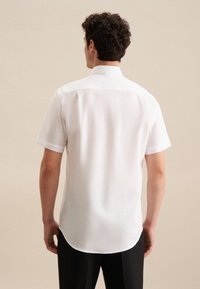Korte mouwen witte blouse met een rechte kraag, gladde textuur en afgeronde zoom, gedragen door een persoon van achteren tegen een neutrale achtergrond.