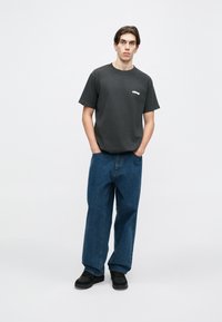 T-shirt de algodão cinza escuro com um pequeno logo branco, combinada com jeans azuis largos e sapatos pretos, de pé contra um fundo branco.