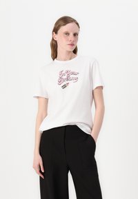 Tricou alb din bumbac, cu text roz "In Your Dreams", cu guler rotund și mâneci scurte, purtat cu pantaloni negri.
