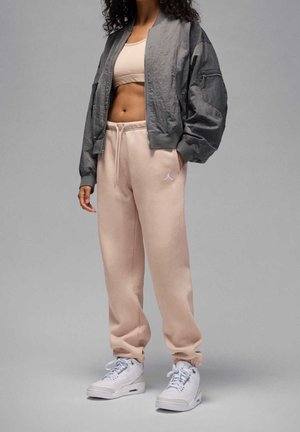 Vrouw met lichtroze joggingbroek, beige sportbeha, grijze bomberjas en witte sneakers, staand op een grijze achtergrond.