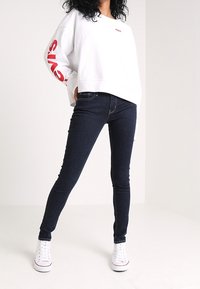 Sudadera blanca con texto "Levi's" en rojo en las mangas, combinada con unos jeans ajustados azul oscuro y zapatillas blancas. Diseño de atuendo simple y casual.
