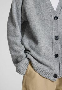 Šedý cardigan vyrobený z měkkého materiálu, s hlubokým V-výstřihem, žebrovanými manžetami a lemem a černými knoflíky. Spárováno s khaki kalhotami.