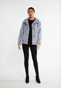 Sær grey jakke med stor krave og to forlommer, prydet med tre guldknapper og en blød tekstur. Båret over en sort top og leggins.