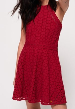 Femme portant une robe rouge en dentelle sans manches avec une taille cintrée et une jupe évasée, debout devant un fond uni.