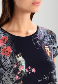 Kvinna i mörkblå skjorta med blommiga och uggletryck, inklusive röda och lila blommor samt paljetterade uggledesigner nära halsringningen.