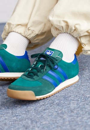 Personne portant des baskets Adidas bleu canard et bleu avec des chaussettes blanches et un pantalon beige sur un sol texturé gris.