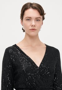 Vestido negro de lentejuelas con escote en V profundo y mangas largas. Presenta una cintura ajustada y una textura suave con detalles reflectantes.