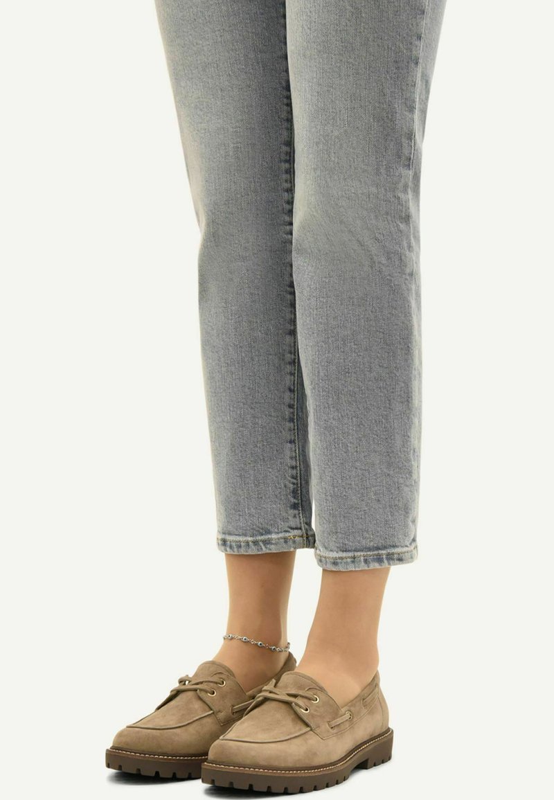 Personne portant un jean court bleu clair, des mocassins marron à lacets avec des semelles épaisses, et un bracelet de cheville argenté à la cheville gauche.