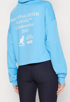 Person iført lyseblå cropped hoodie med hvid tekst og logoer på ryggen, kombineret med mørkeblå tætsiddende jeans.