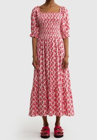 Kukallinen maxi-mekko, jossa on neliönmuotoinen pääntie, puffihihat, smockattua vartalonosa ja kerroksellinen hameosa, pinkissä värissä punaisilla kukilla. Yhdistetty pinkkeihin sandaaleihin.