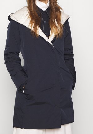 Žena oblečená v námořnicky modrém kabátu s bílým lemováním kapuce, s rukama v kapsách, přes černou košili na knoflíky a bílý rolák.