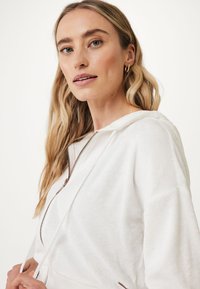 Felpa bianca con zip realizzata in morbido tessuto di spugna, dotata di chiusura frontale con zip, cordini e una vestibilità rilassata con dettagli a costine sul bordo.