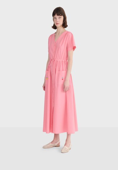 Femme portant une robe maxi rose à boutons sur le devant, manches courtes, taille à cordon, poches avant avec une petite broderie colorée, et des chaussures beige à enfiler.