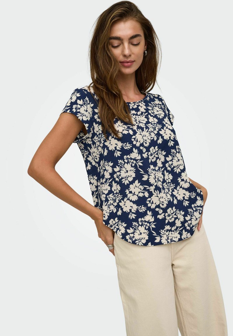 Femme portant un haut bleu marine à manches courtes avec un motif floral blanc et un pantalon beige clair, debout les mains dans les poches.