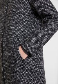 Cappotto texturizzato in bianco e nero con zip frontale, dotato di tasca laterale e silhouette dritta, realizzato in tessuto intrecciato.