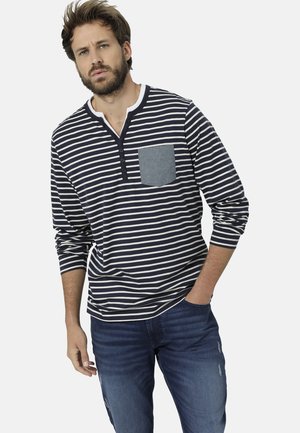Gestreiftes Langarmshirt in Marineblau und Weiß mit einer grauen Brusttasche; verfügt über einen Henley-Ausschnitt und weichen Stoff. Blaue Jeans mit leichten Used-Effekten.