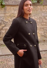 Cappotto di lana nero con design doppiopetto, bottoni dorati ornamentali, tasca con hardware dorato e vestibilità sartoriale.